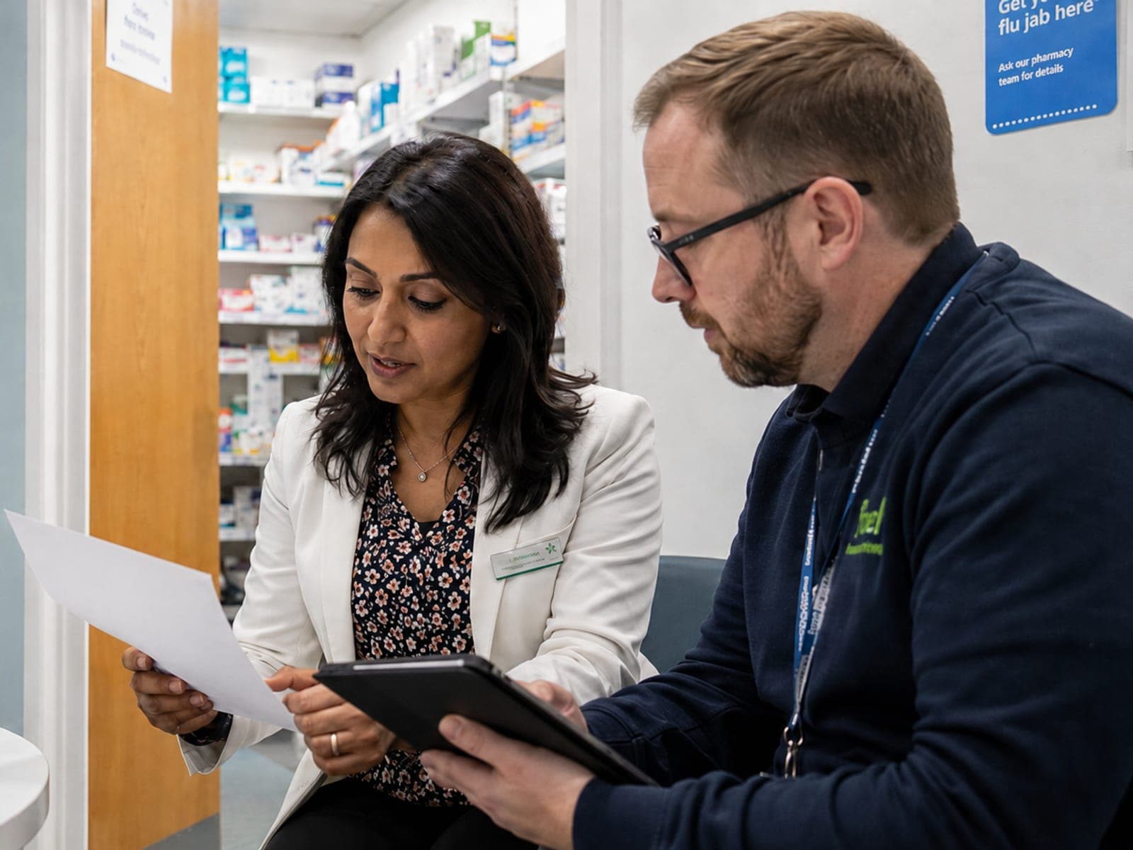 Two community pharmacists reviewing a document together in the dispensary, with NHS pharmacy shelving and signage behind them